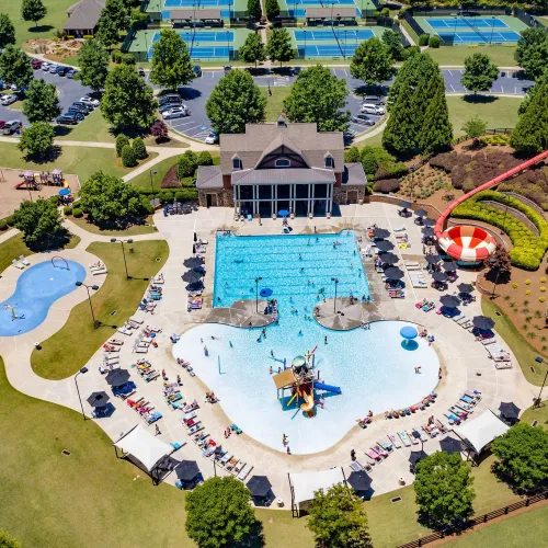 Aerial view of a community pool area with slides, splash pads, tennis courts, playground, and surrounding green spaces.